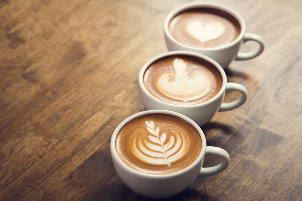 Three cups of latte coffee with intricate latte art designs arranged on a rustic wooden table in a cozy café setting.