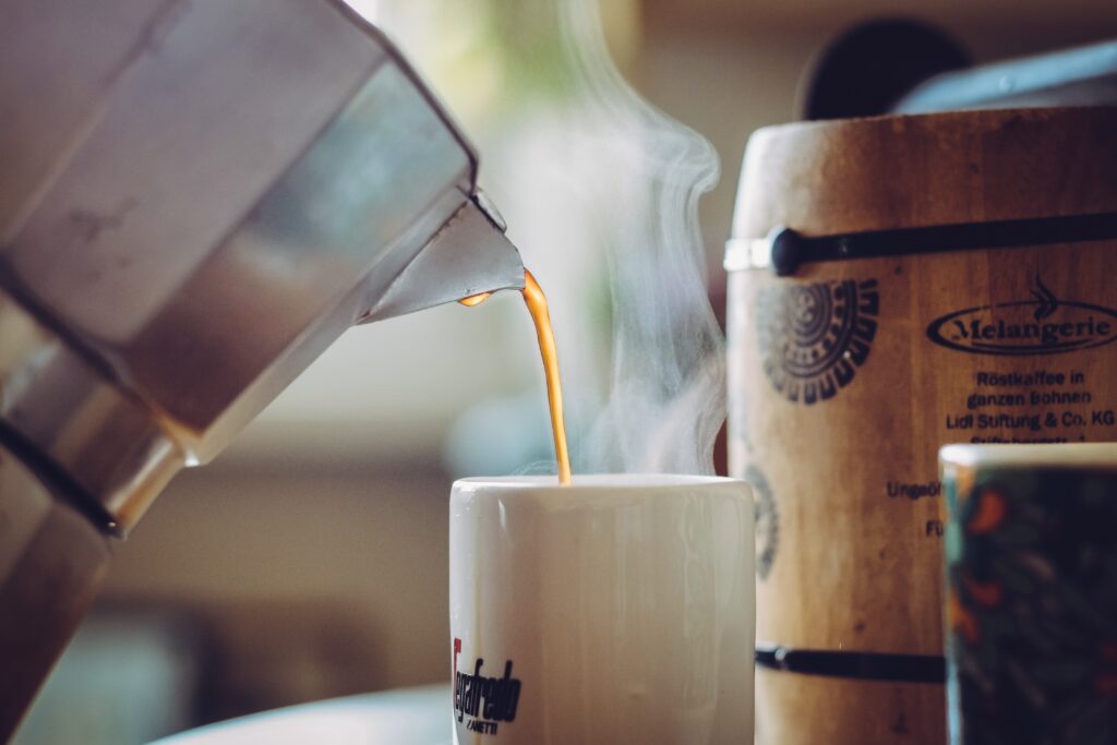 Fresh hot coffee being poured from a stovetop moka pot into a white ceramic mug with visible steam rising in a cozy kitchen setting.