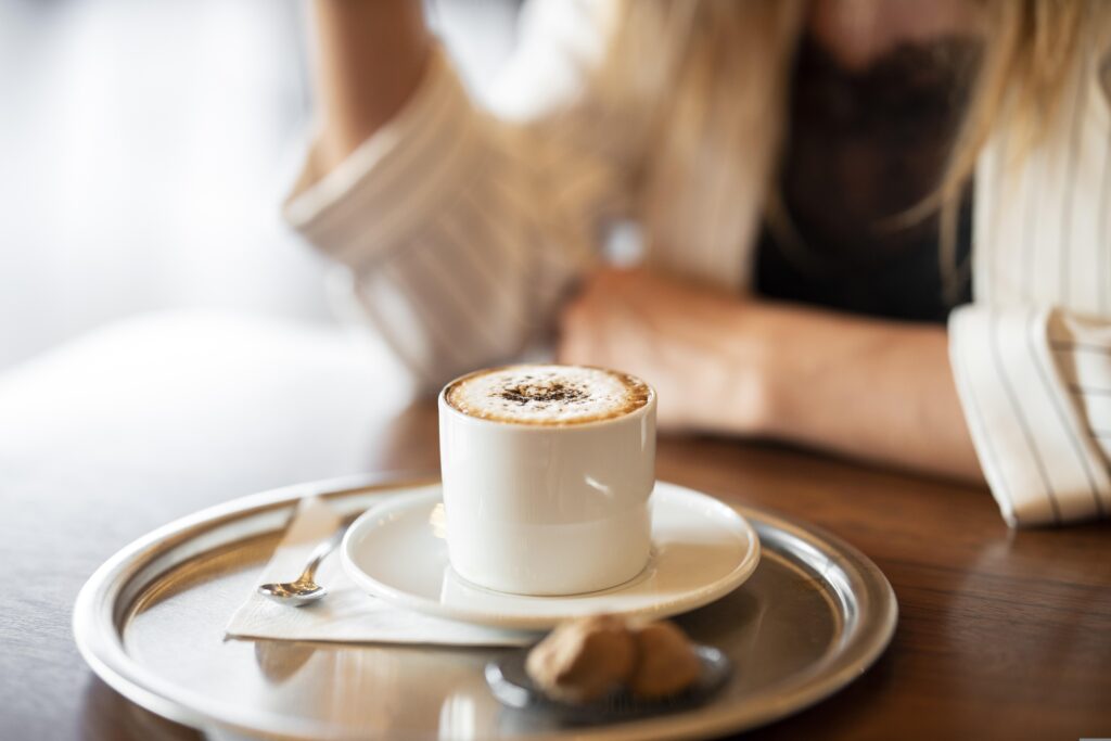 Fresh cappuccino served in a white cup on a tray with a spoon and cookies, placed on a wooden café table with a blurred person in the background.
